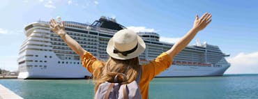 A backpack-clad traveller stands on a dock facing a large white cruise ship, arms raised in excitement under a clear blue sky.