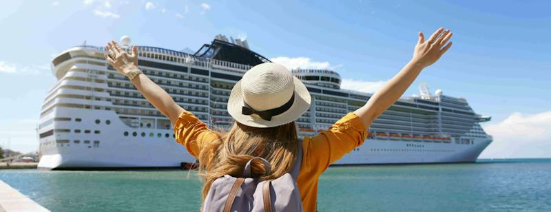 A backpack-clad traveller stands on a dock facing a large white cruise ship, arms raised in excitement under a clear blue sky.