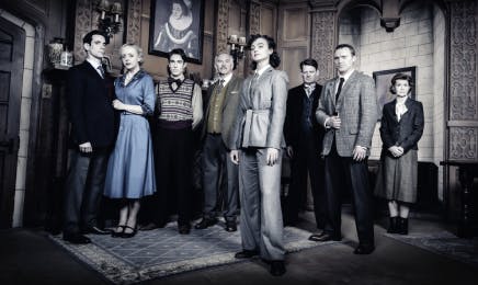 The cast of 'The Mousetrap' standing in a line on stage, dressed in period costumes against the backdrop of the play's set, all with serious expressions indicative of the mystery's high stakes.
