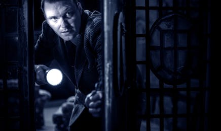 In a tense scene from 'The Mousetrap,' an actor illuminated in blue light searches through a dark room with a flashlight, peering through a window with an expression of concentration and urgency.