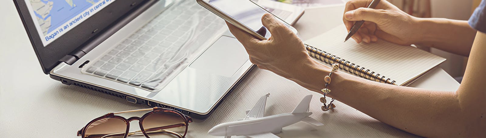 Person planning a trip at a desk with a laptop, phone and notebook, next to a toy plane and sunglasses.