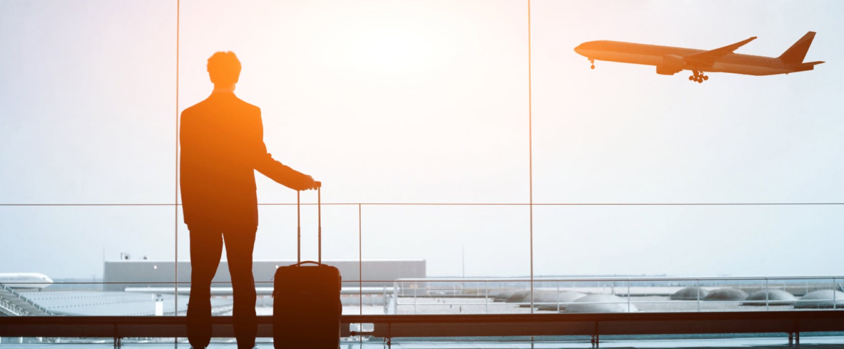 Person standing in an airport terminal with a suitcase, watching a plane take off at sunrise through large glass windows.