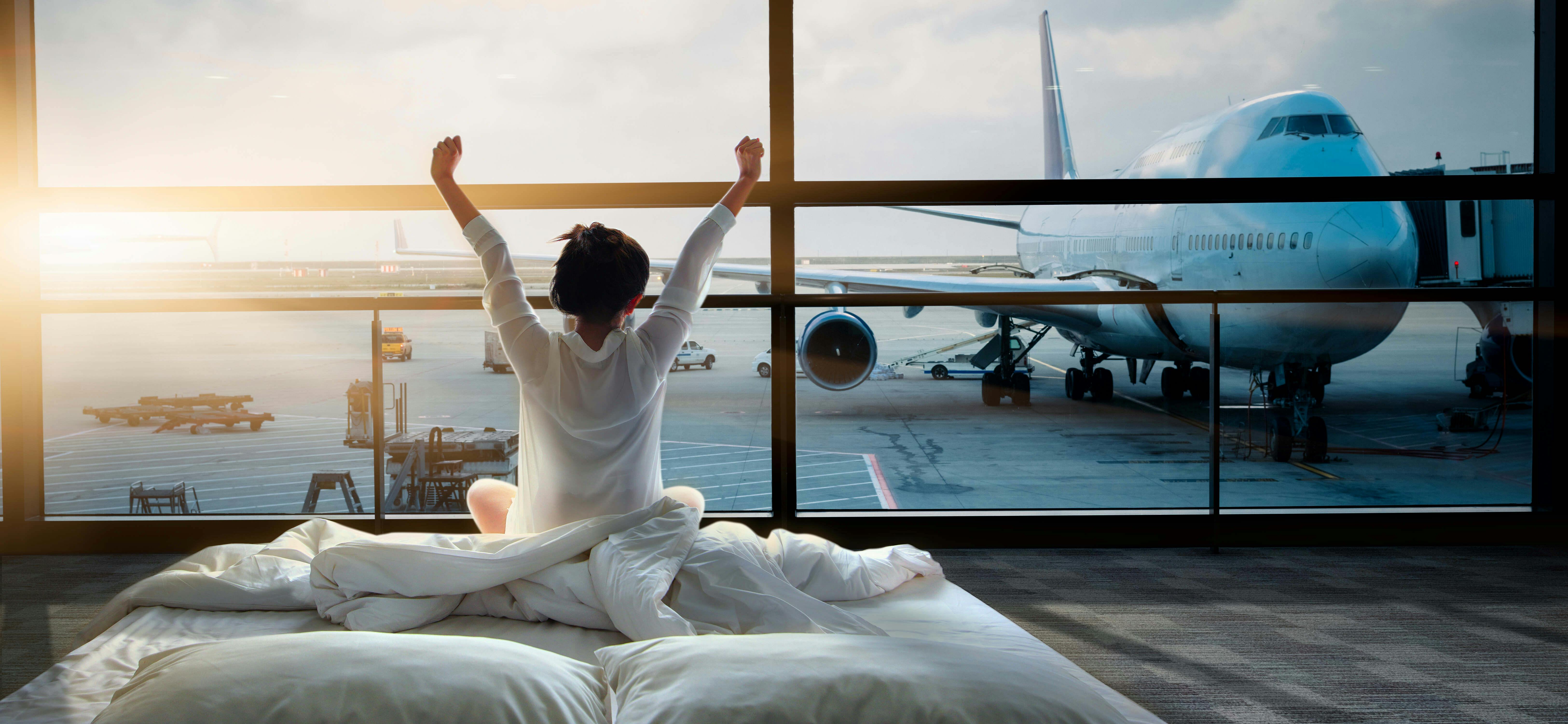A guest in a white bathrobe sits up in bed at an airport-side hotel room, stretching their arms toward a floor-to-ceiling window that looks out over a parked airliner at sunrise.