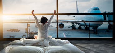 A guest in a white bathrobe sits up in bed at an airport-side hotel room, stretching their arms toward a floor-to-ceiling window that looks out over a parked airliner at sunrise.
