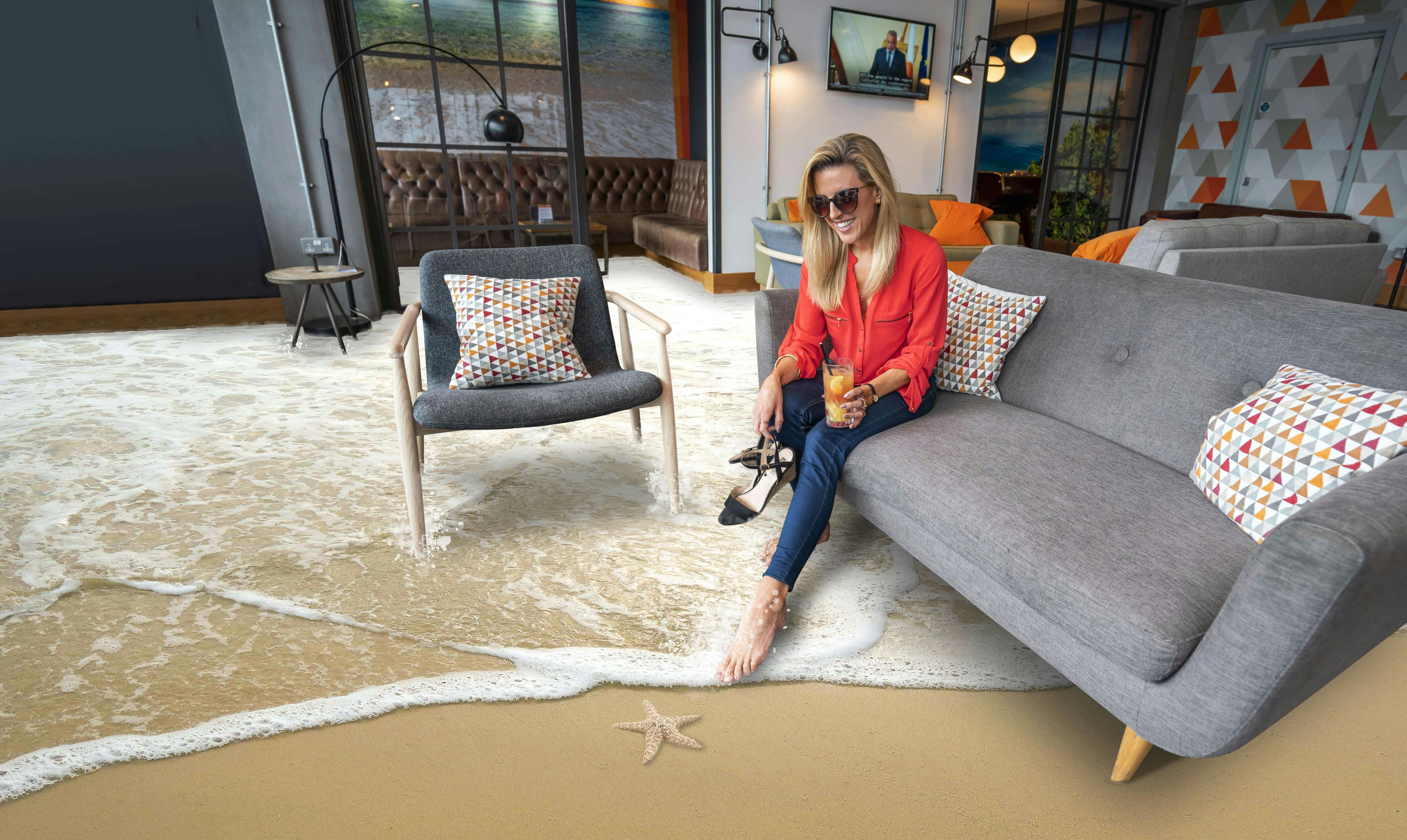 A woman in a red blouse and sunglasses sits on a grey sofa in a living room where water has flooded the carpet, dipping one bare foot into the shallow pool while holding her shoes and a drink.