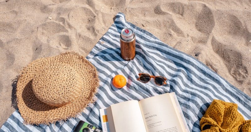 An image of a sun hat, camera, sunglasses and a book on a beach towel