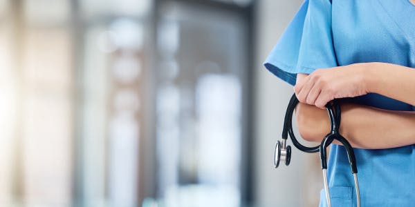 Close-up of a healthcare professional wearing blue scrubs, holding a stethoscope in crossed arms. The background is blurred, suggesting a modern, well-lit medical facility or hospital setting.