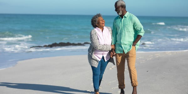 An older couple walking barefoot along a sandy beach, holding hands and smiling at each other. The man is wearing a light green shirt and tan trousers, while the woman is dressed in a light cardigan and pink blouse. The ocean waves gently crash in the background under a clear blue sky.