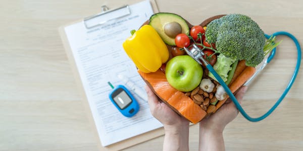 Hands holding a heart-shaped bowl of cholesterol-friendly foods—broccoli, avocado, yellow pepper, cherry tomatoes, apple, salmon and almonds—wrapped in a stethoscope, set beside a medical form and a glucose meter, illustrating travelling with high cholesterol.