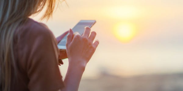 A person standing on a sandy beach, holding and tapping a smartphone, with the sun setting over the ocean and a golden sky in the background.