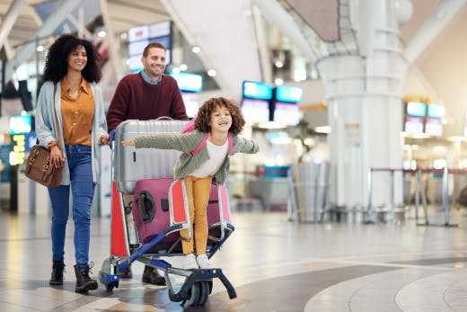 miling family walking through an airport; a young girl rides on a luggage trolley with arms outstretched while her parents walk beside her.