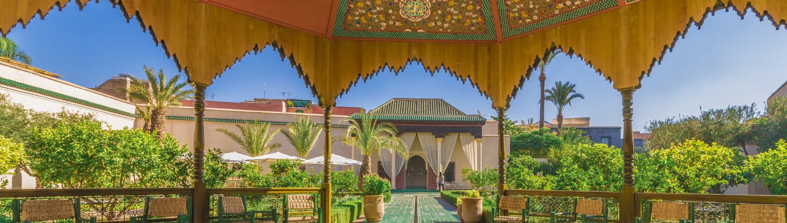 A panoramic view from beneath an ornately carved wooden gazebo in a lush, sunny Moroccan Riad courtyard.