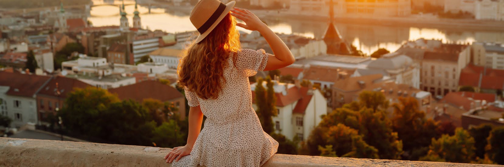 A woman in a sunhat and light summer dress sits on a ledge overlooking a city at sunset, admiring the view of rooftops, a river, and distant bridges.