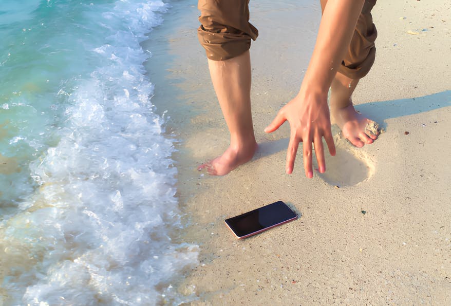 Person on a sandy beach reaching for a smartphone dropped near the shoreline as waves approach.