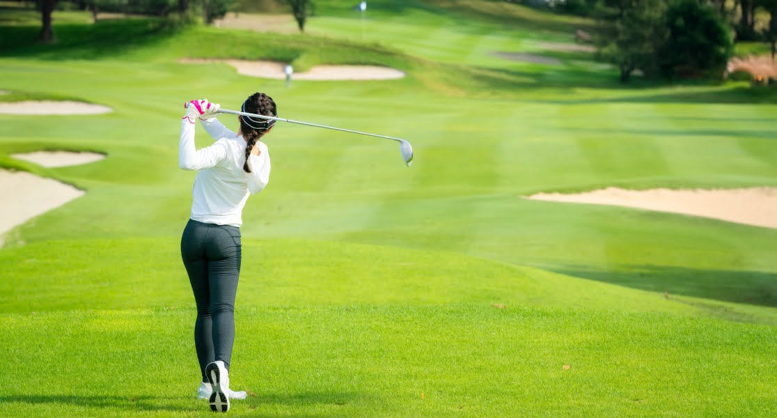 A woman swinging a golf club on a bright green course under clear skies, representing outdoor sports and leisure while travelling.