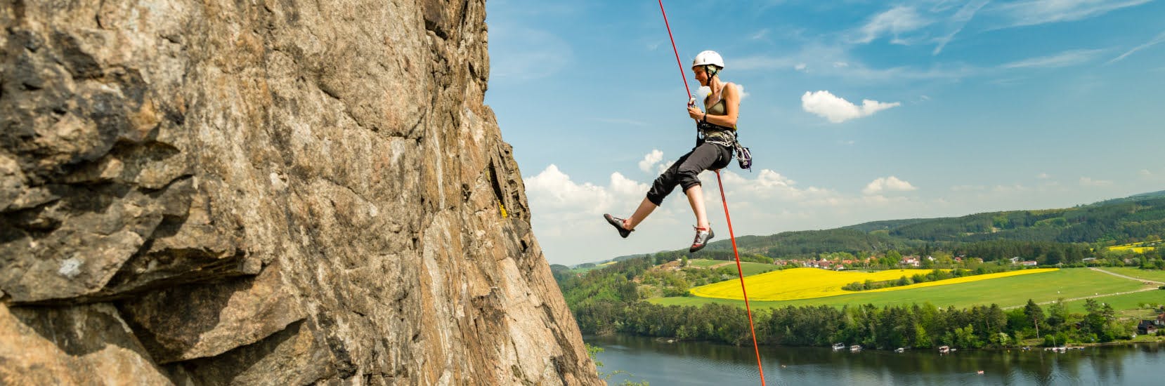 Person abseiling down a rocky cliff face with a river and countryside in the background on a sunny day