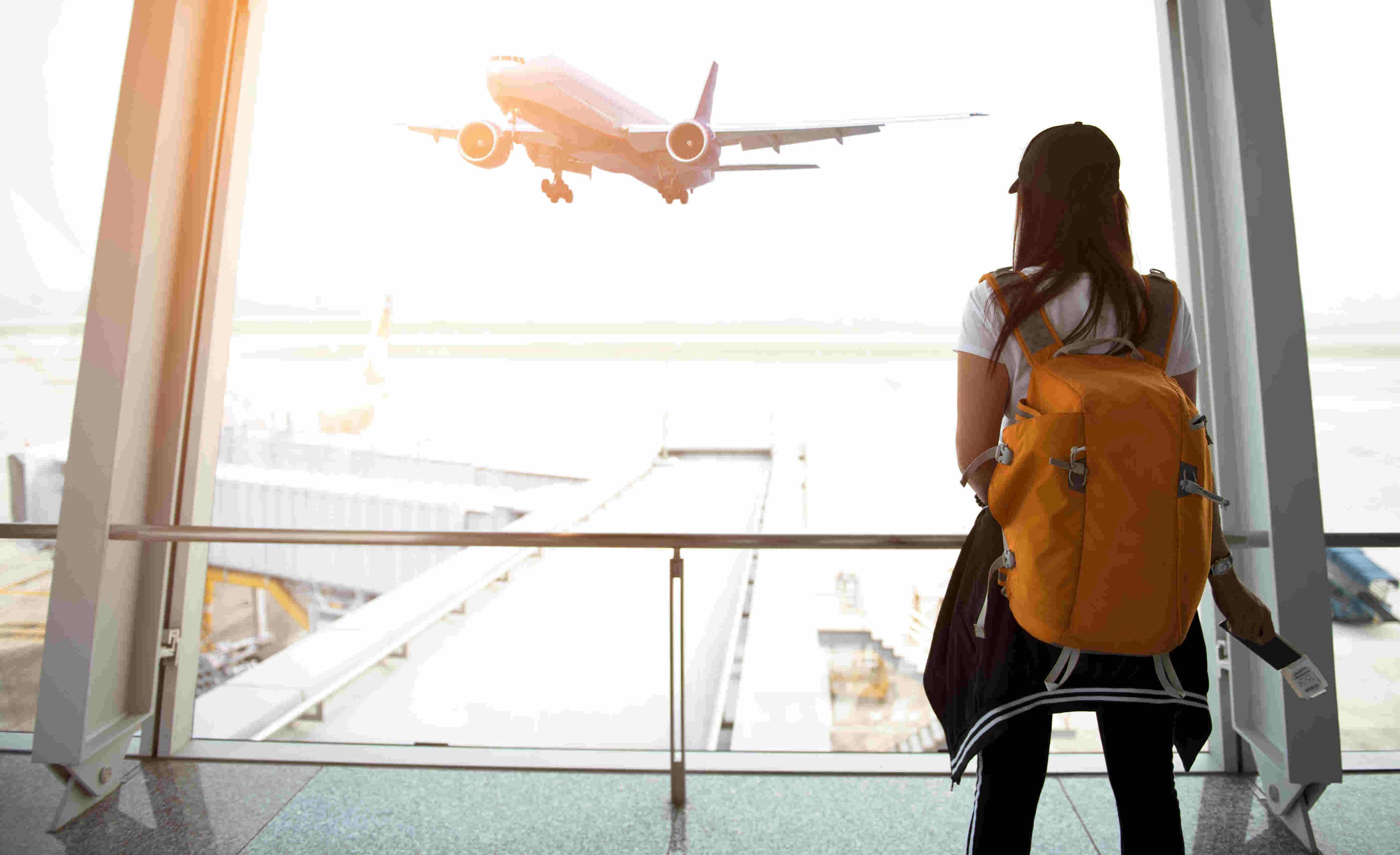A woman with a bright orange backpack stands at an airport terminal window, holding a passport and boarding pass, watching a plane take off into the sky.