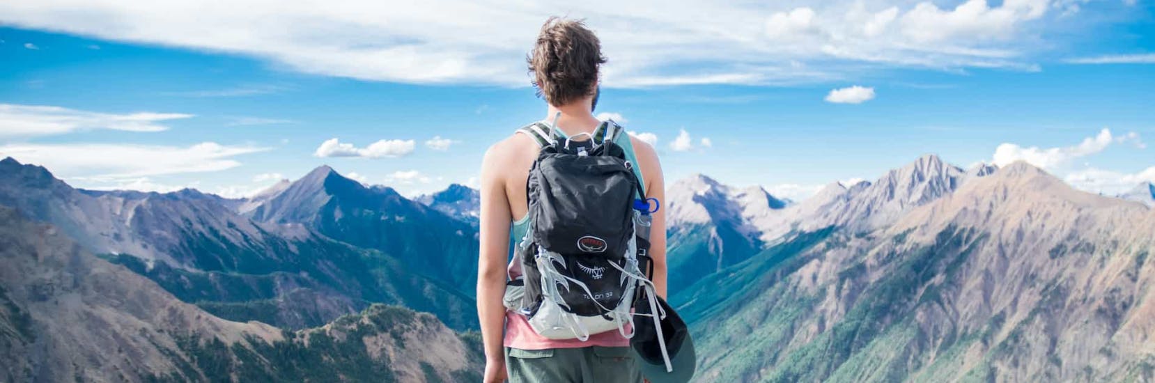 Backpacker standing on a mountain ridge overlooking vast peaks and valleys under a bright blue sky.