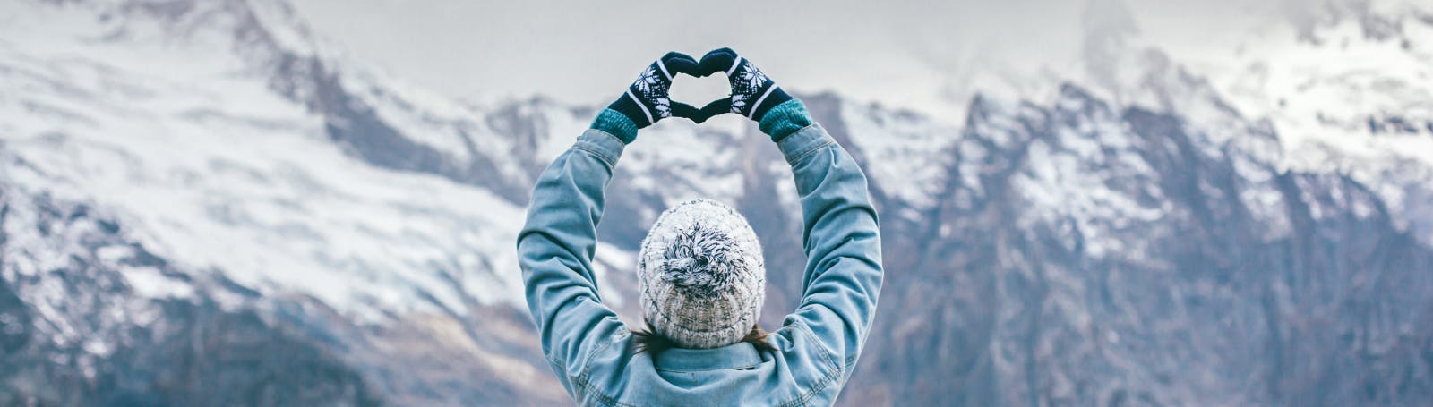 Person standing in front of snow-covered mountains forming a heart shape with their hands, symbolising love and care.