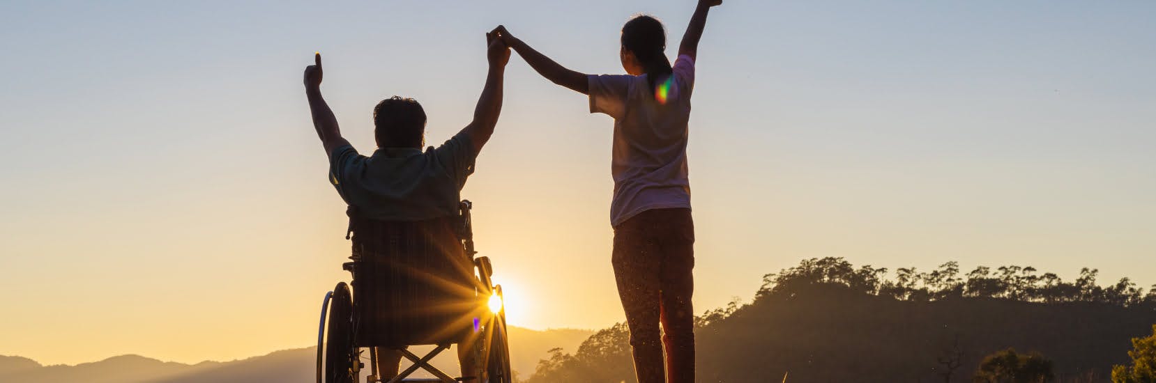 A person in a wheelchair and their companion hold hands and raise their arms as the sun sets over distant hills.