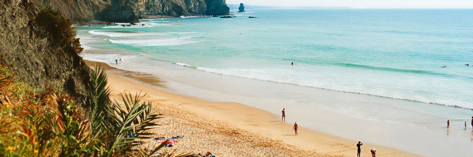 Golden sandy beach surrounded by cliffs in the Algarve, Portugal, with a few people walking and swimming in the clear turquoise sea under a bright sky.