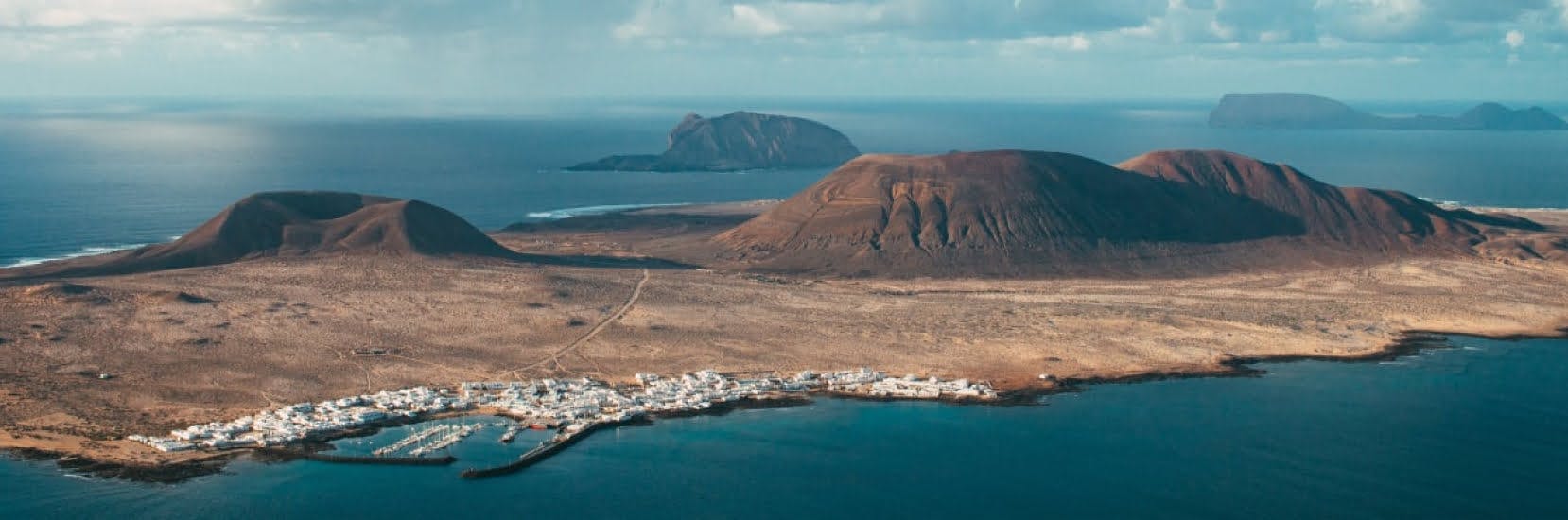 Volcanic landscape of La Graciosa island in the Canary Islands, Spain, with rugged hills, a small coastal village, and the deep blue Atlantic Ocean in the background.