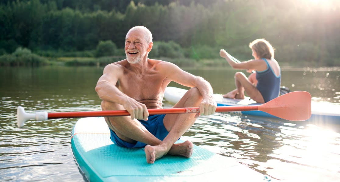 Older couple paddleboarding on a serene lake, enjoying an active holiday with the reassurance of annual travel insurance