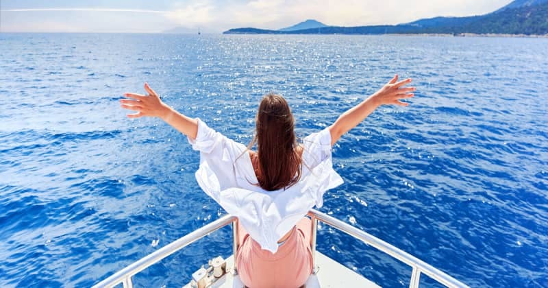 A woman with outstretched arms enjoys the fresh sea breeze while standing at the front of a boat