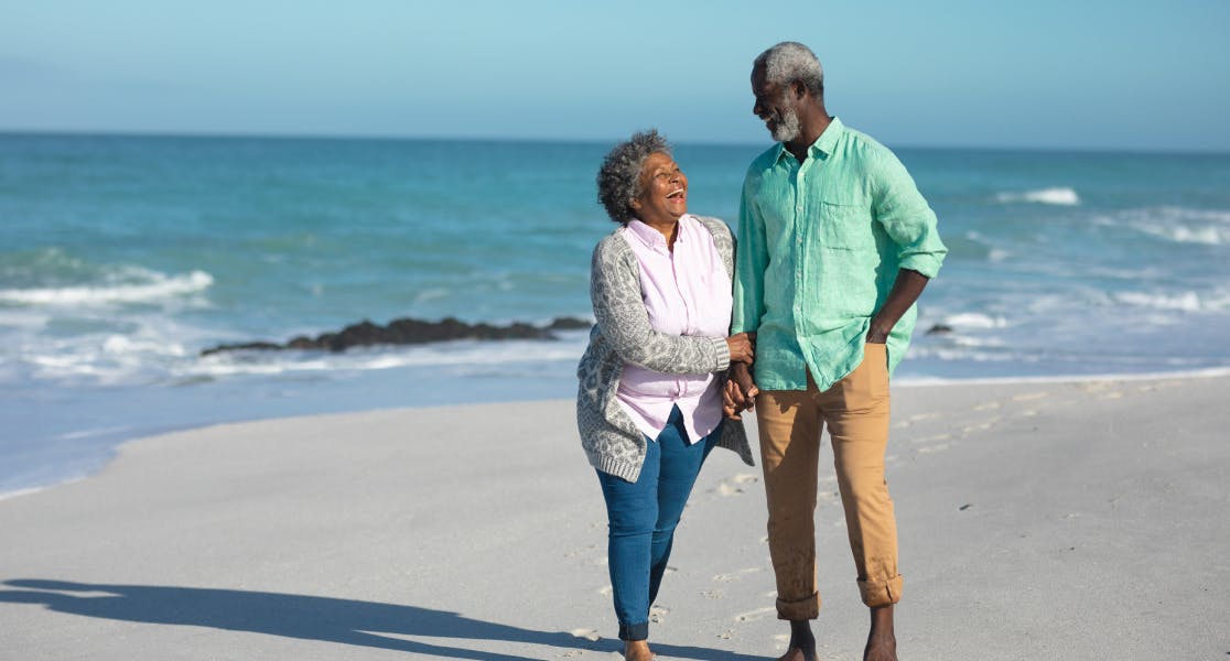 An older couple walking barefoot along a sandy beach, holding hands and smiling at each other. The man is wearing a light green shirt and tan trousers, while the woman is dressed in a light cardigan and pink blouse. The ocean waves gently crash in the background under a clear blue sky.