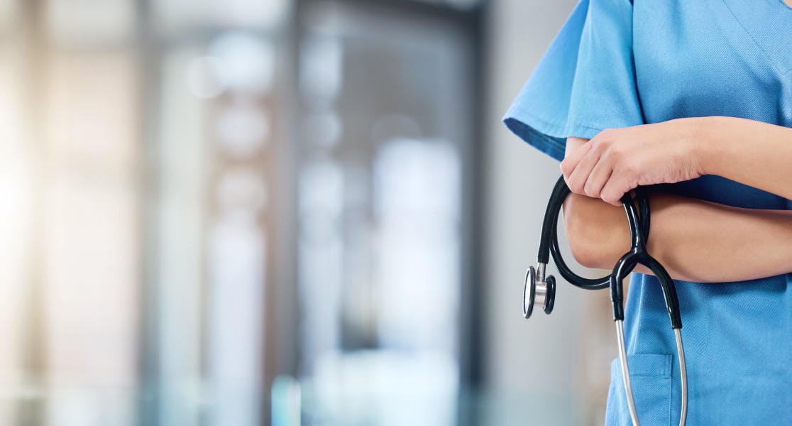 Close-up of a healthcare professional wearing blue scrubs, holding a stethoscope in crossed arms. The background is blurred, suggesting a modern, well-lit medical facility or hospital setting.