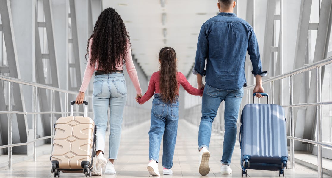 Back view of a family walking through an airport terminal. The parents, each pulling a suitcase, hold hands with their young daughter in the middle. They are casually dressed in denim outfits, and the setting appears to be a modern, well-lit hallway with large windows