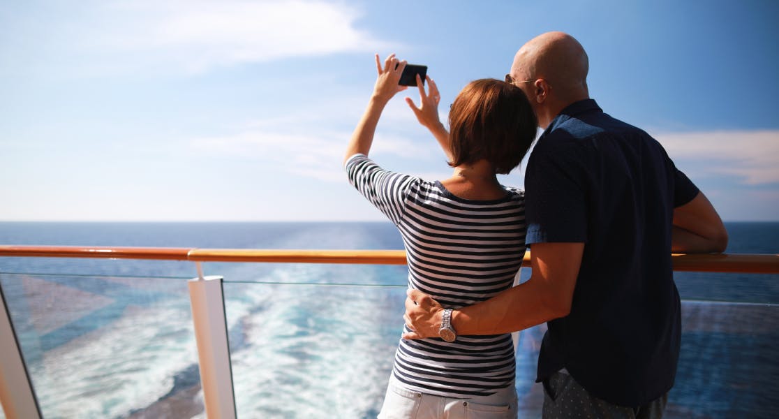 Middle aged couple on a cruise ship looking out to a blue ocean and sunshine. The lady holds a phone up, taking a picture of the view.