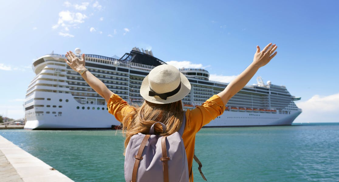 A woman standing on a pier with her arms raised in excitement, facing a large cruise ship docked nearby. She is wearing a straw hat, a yellow shirt, and a backpack, with a bright blue sky and calm waters surrounding the scene.