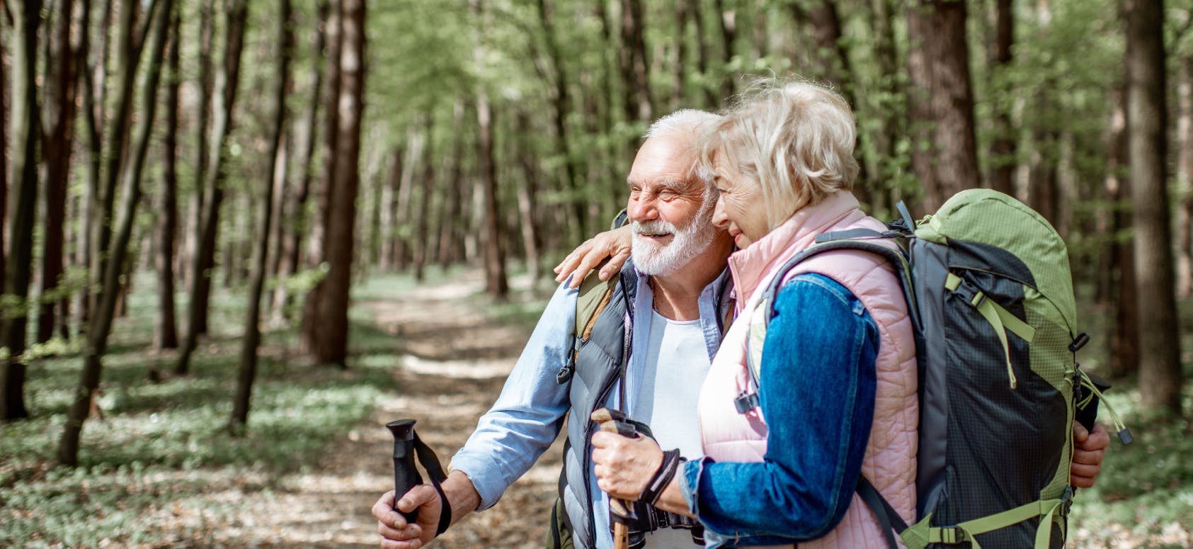 Elderly couple hiking in a forest, smiling and embracing while carrying backpacks and walking sticks.