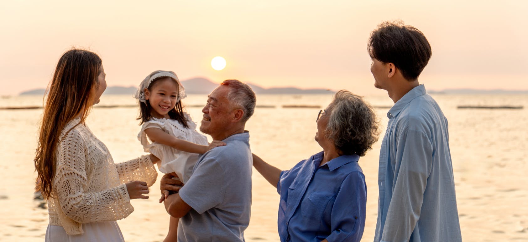 Smiling multigenerational family standing on the beach at sunset, with a grandfather holding a young girl while the others look on happily