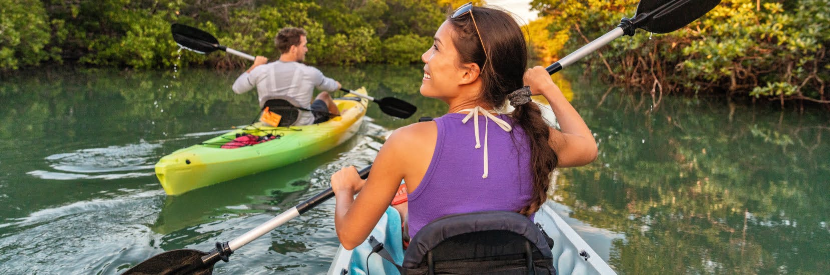 Two people kayaking through calm green water surrounded by lush trees, with a woman in the foreground smiling as she paddles and a man kayaking ahead of her.