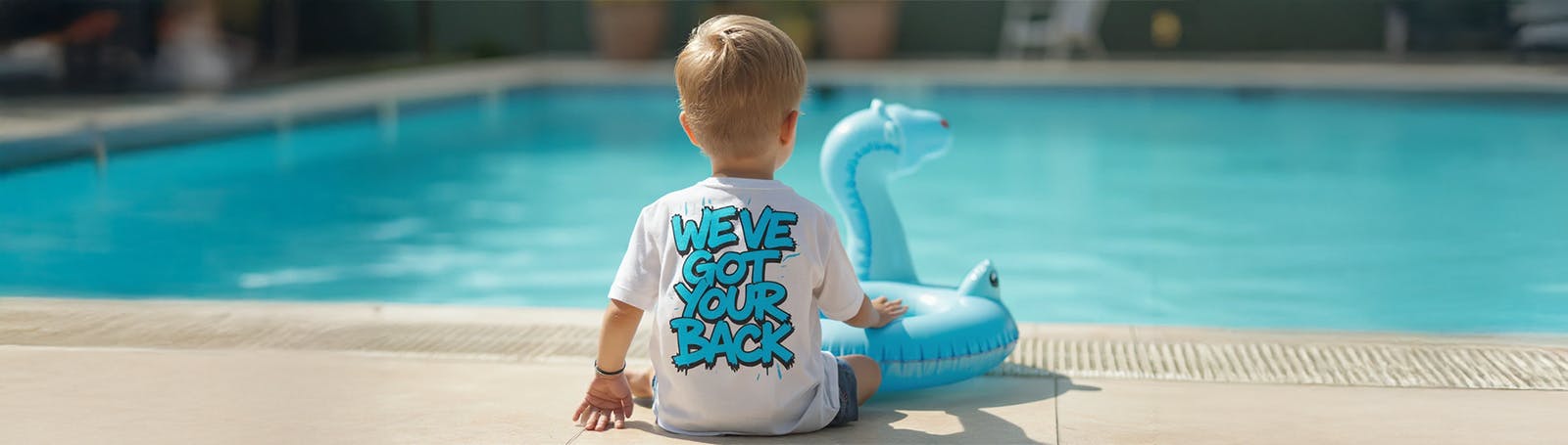 A young child sits at the edge of a swimming pool with their back to the camera, wearing a white T-shirt that says 