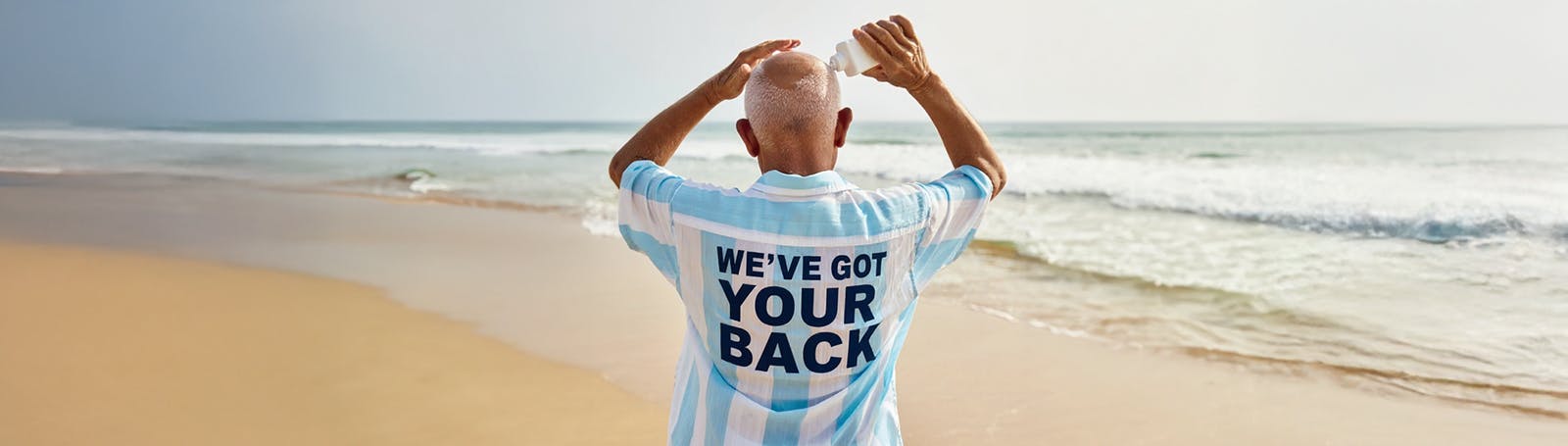 An older man stands on a sandy beach with his back to the camera, wearing a light blue striped shirt that reads 