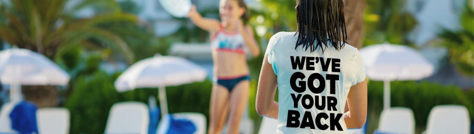 A parent with wet hair stands at the edge of a sunny pool wearing a T-shirt that says 
