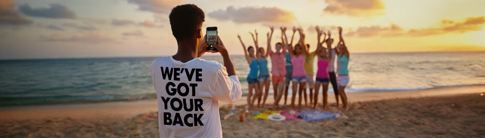 A young person stands on the beach at sunset, taking a photo of a group of friends who are posing with their arms raised. The photographer wears a T-shirt with the words 