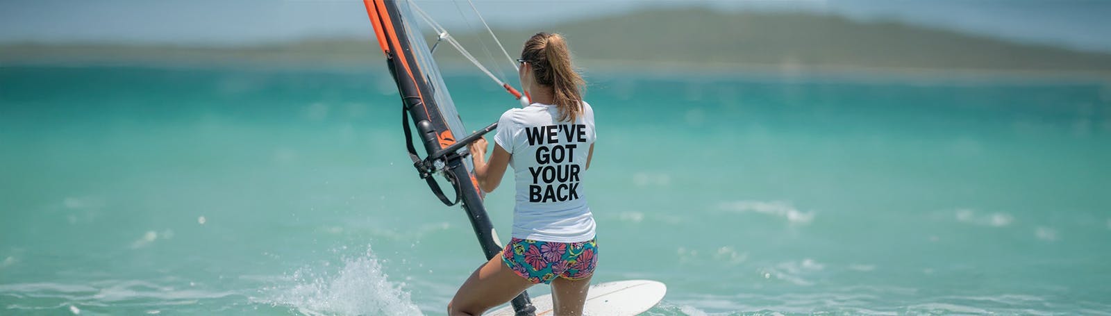 A woman windsurfs in bright turquoise water, seen from behind as she holds the sail and balances on the board. She wears a white T-shirt with the words ��We've Got Your Back