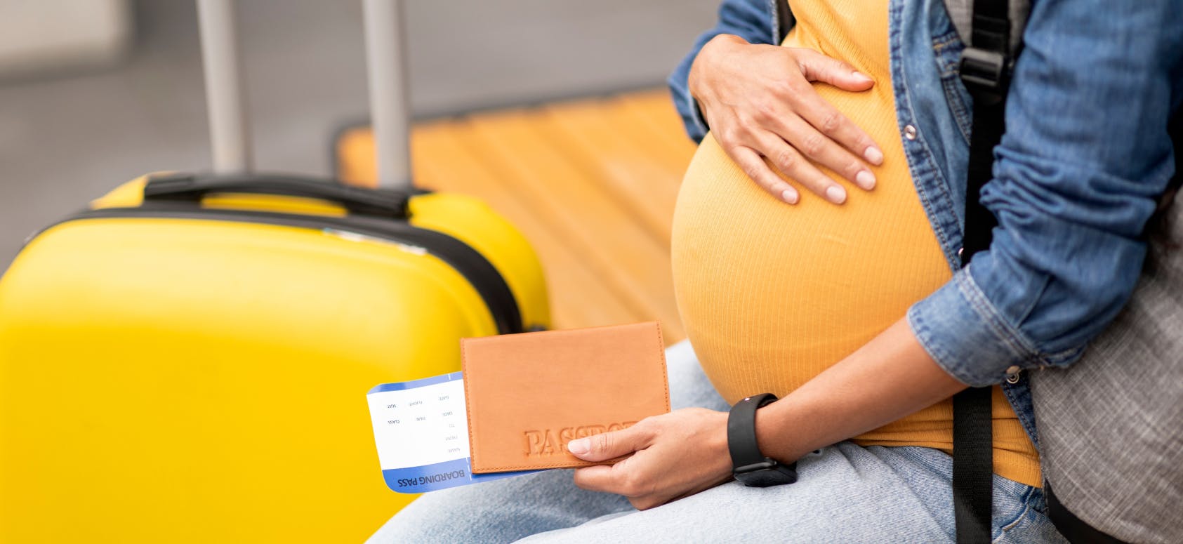 Pregnant traveller at an airport holding a passport and boarding pass beside a yellow suitcase.