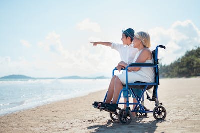 Urlaub mit Rollstuhl: Sohn und Mutter im Rollstuhl blicken am Strand in die Ferne.