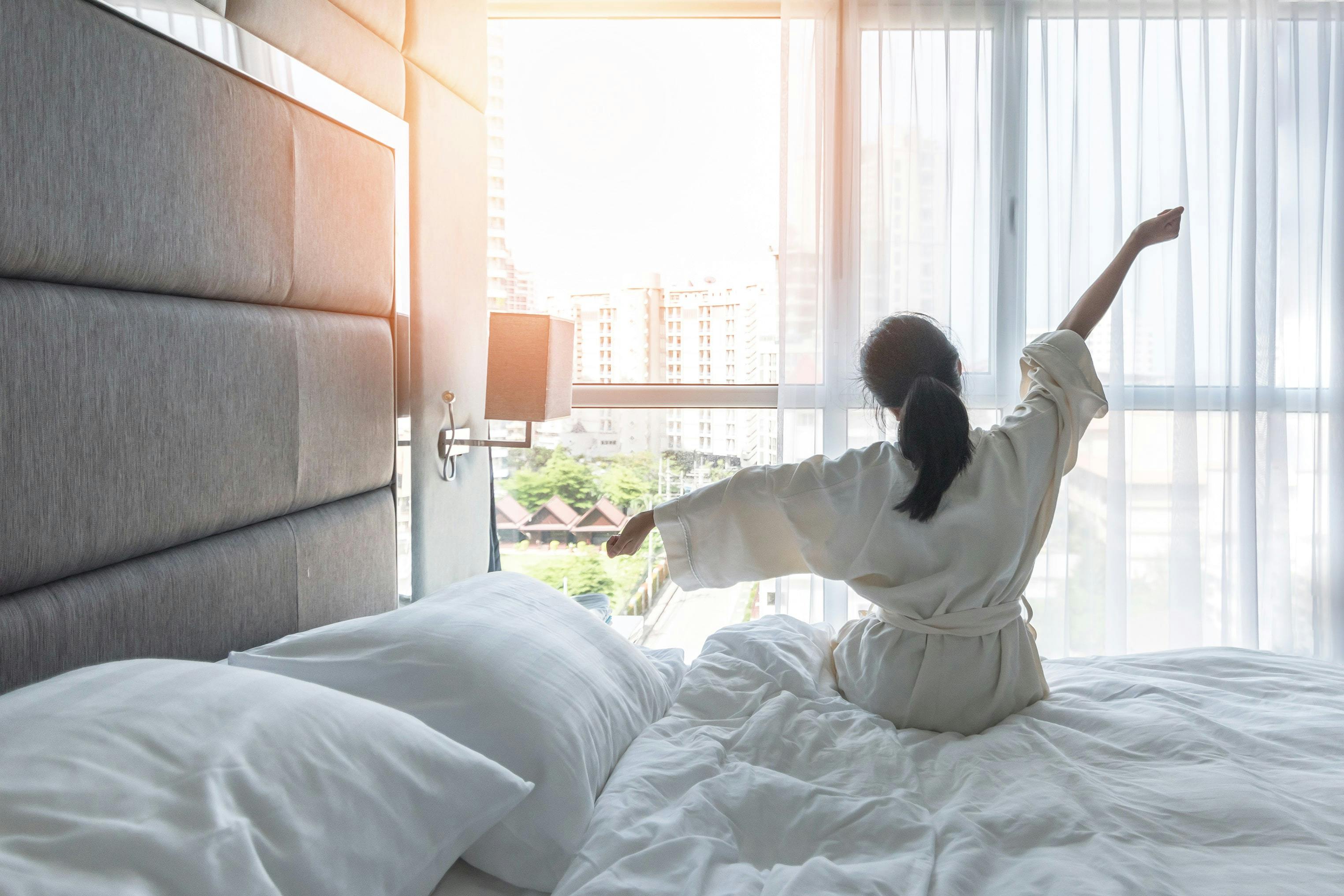 A woman in a white bathrobe sits up in a hotel bed, stretching her arms toward sunlit floor-to-ceiling windows overlooking a city.